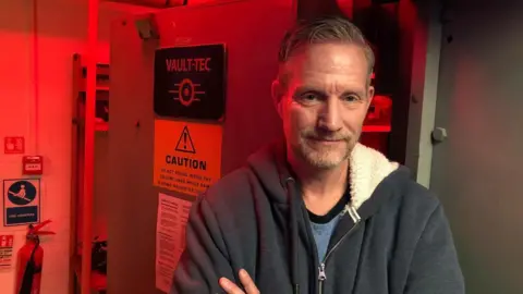 Vikki Irwin/BBC Jon Betts is standing in front of a bank vault door. He is wearing a blue hooded top and has his arms folded. 