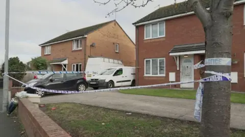 Police cordon around the front garden of a house, with vehicles in the drive, and police tape tied to a tree and lamppost