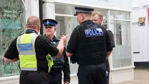 Four male police officers in uniform are talking to each other in a street with shops behind them. One with his back to the camera is wearing a hi-visibility waistcoat with 'Community Safety Team' printed on the back and the other is wearing a stab vest that has 'Police' printed on the back. 