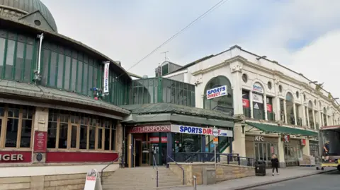 The shopping centre. There is a red pub to the left of the image and a number of shops inside the shopping centre on the right.