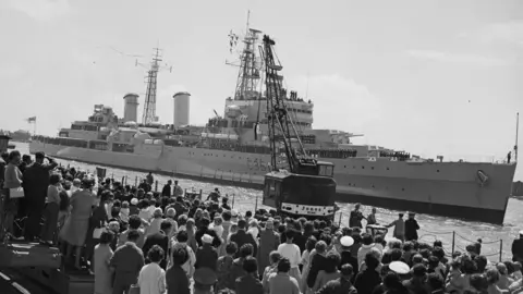 Getty Images A black and white picture from the 1960s of a warship arriving in port. There are hundreds of people on the dock watching the ship return.