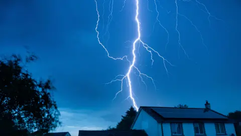 Cumbrian stargazer A dark blue sky above a detached house with a bright, large lightening bolt in the centre. The lightening has one strong line down the centre, with small offshoots which look like veins.