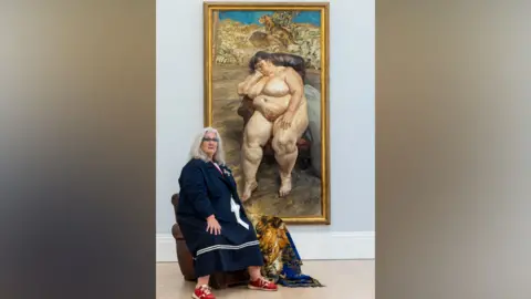 Sotheby's Sue Tilley is sitting on a chair in a gallery beside a large framed painting of a reclining nude figure. The gallery has a light grey wall, wooden floor, and a patterned blanket draped over the chair.