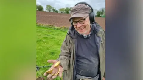 Johnathan is holding the item in his right hand as he stands in a field. He is wearing a scruffy jumper, jacket and cap. He is also wearing glasses. 