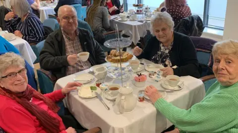 Saltbox Four elderly people sit around a dinner table drinking tea and eating cake.