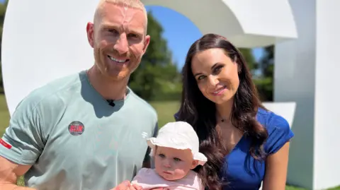 A man in a pale blue T-shirt and a woman in a blue top hold a baby between them which is wearing a pink outfit and hat