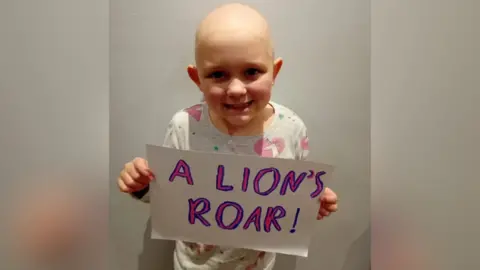 Cancer Research UK Lily holding a sign that says "a lion's roar" in pink and blue writing.