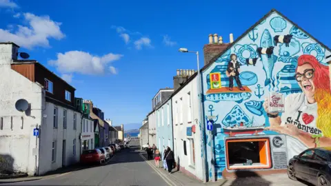 A narrow street in Stranraer with people walking down it and a colourful mural celebrating the town on the right hand side