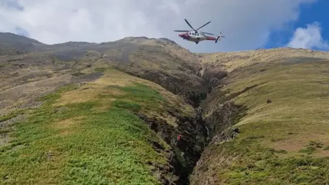 A red helicopter flies low in the Wasdale Valley above Ill Gill, Kirk Fell. The gill is steep, black and runs very deep. A mountain rescue volunteer is being winched by the helicopter over the ravine. There are emergency vehicles in the distance. Grey scree slopes and bracken feature on the green terrain.