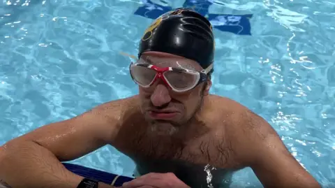 Steve Harris, a man leaning on the edge of a pool wearing goggles and pursing his lips as if he is sighing.