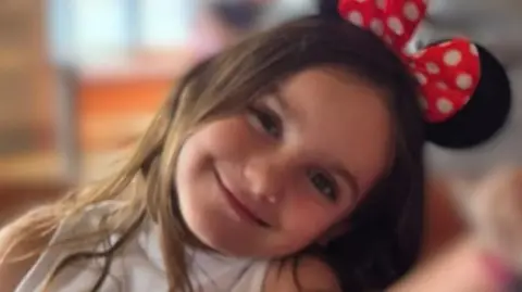 Family Aria, who has long brown hair, smiles at the camera with her head tilted to one side. She is wearing a black pair of Minnie Mouse ears with a red and white polka dot bow on them. 