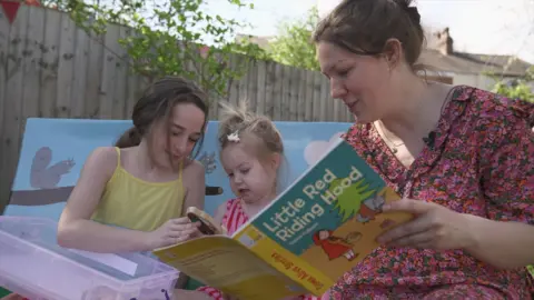 Hannah Karpel / BBC A mum and her two daughters sit on the bench in the library garden as she reads Little red riding hood to them. The youngest toddler sits in the middle holding a wooden wolf from the story.