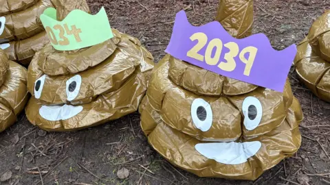 A close-up of two brown inflatable poo swirl emojis with coloured hats, which show the years 2034 and 2039. They have wide eyes and mouths. They are standing on mud and grass.