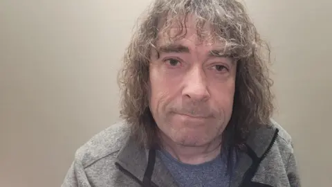 Matthew Wildman A man with long curly hair in his 50s.
