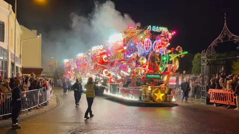 A multi-coloured illuminated cart is making its way through a town centre. Crowds of people are standing behind barriers and taking pictures.