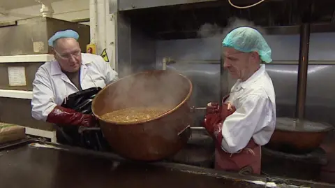 Two factory workers wearing white coats, leather aprons and gloves stand are holding a big copper pot. Inside the pot is orange bubbling liquid which is being poured out onto a brown cooling slab. 
