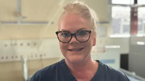 A women in her 50s with tied back blonde hair stands facing the camera and smiling. She is wearing glasses with a tortoise shell frame and a dark blue top. She s stood in a room with hospital apparatus behind her.