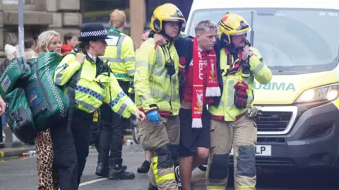 PA Media Jack Trotter, with blond hair and with a red Liverpool scarf around his neck, is helped by two fire officers and a police officer in the aftermath of the incident.