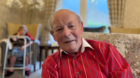 A man sat in a chair wearing a red shirt with white, green and blue thin vertical stripes on it and a white collar. Top button undone. Blurred background shows a beige room. 