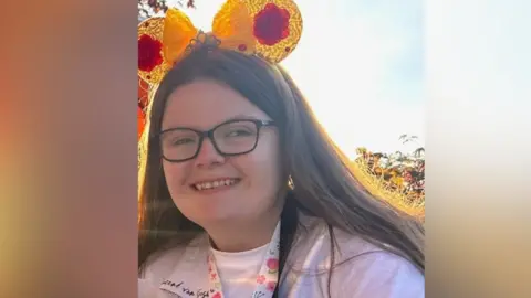 A woman with glasses and wearing a tiara smiles to the camera. She is sitting outside.