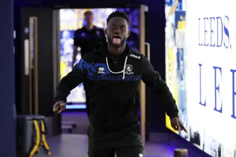 Tom Banks/MFC A footballer in a black tracksuit sticks his tongue out as he walks towards the camera next to a large illuminated sign that reads "Leeds".