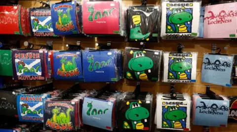 Getty Images A wall of t-shirts in plastic hanging on rails. The t-shirts have different images of the Loch Ness Monster and are different colours. Most of the images show Nessie with three humps.