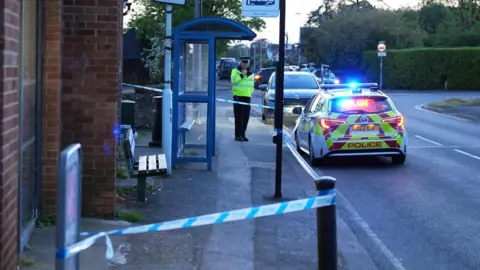 A police officer stands next to a police car with flashing lights on a cordoned off section of road. 