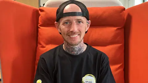 Steven wearing a black baseball cap and a black t-shirt, with an owl tattoo on his neck.