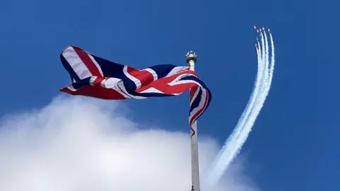 The Union Flag flies from a flagpole. In the distance, the Red Arrows aircraft in formation trail white smoke in a blue sky 