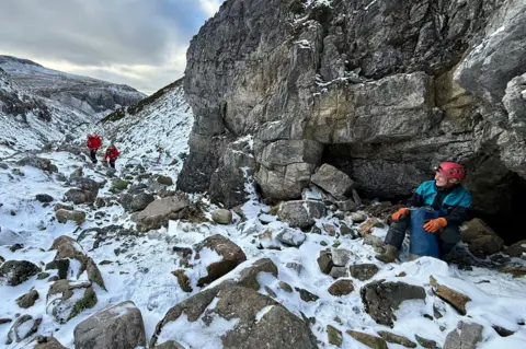 Assynt Mountain Rescue Team A rescue volunteer sits at the entrance to the cave system. It is in an area of steep, rocky terrain with a dusting of snow. Two other rescue team members are just visible walking along a track.