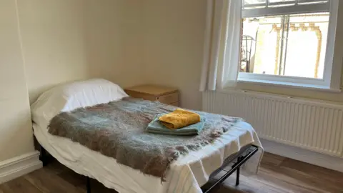Clare Worden/BBC A small tidy bedroom with a single bed. On it is a green coloured blanket and two towels. One is green and one is yellow. 