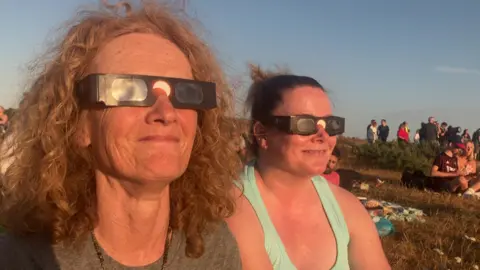 Two women wearing eclipse glasses at Hengistbury Head in Dorset.