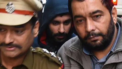 Jagtar Singh Johal being escorted to a court in Ludhiana on 24 November 2017. He is wearing a dark blue hooded top with the hood up. Two men are in the foreground, one in a uniform and ther othey in a grey jacket, and there are a number of people in the background.