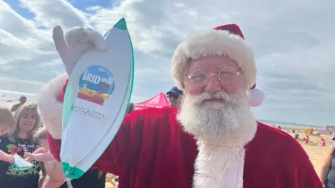 A man dressed in a Santa outfit with a red coat, white fur-style lining and a red hat stands on a beach while holding up what appears to be a goody bag, which is white and green with a "Bridmas Day" logo on it.
