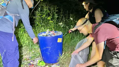 Image of two men and one woman placing litter into an overflowing blue bin and transparent plastic bags in a woodland on the Cotswold Way. 