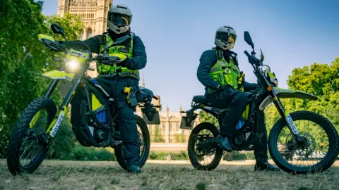 Two Metropolitan Police officers in full riding gear and helmets sit on high-powered Sur-Ron police e-bikes in a park, with the Houses of Parliament visible in the background under a clear blue sky.