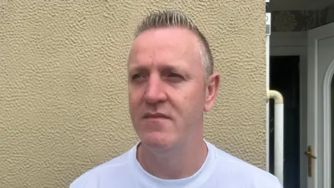 A man with short spiky grey hair wears a white t-shirt as he muses of into the distance. Behind him is beige wall with an open white door over his left shoulder.