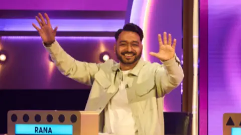 A man with black hair and goatee is wearing a cream-coloured acket over a white tshirt and  sitting behind a brightly lit game show desk on a colourful studio set, raising both hands. The set features purple and pink lighting, geometric patterns and name panels on the desk, giving the scene a lively television game show atmosphere.