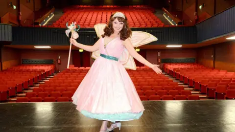 Paul Clapp Melanie Walters standing on stage wearing a pink fairy dress with blue ribbon and petticoat. She is wearing glittery pink wings, a tiara, white heels and is holding a pink wand in the air. Behind her is a theatre with hundreds of red cushioned seats. 