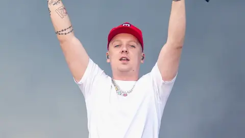 Getty Images Aitch, a male rapper on stage, wearing a red cap, white tshirt with a necklace - with his arms raised high mid performance.