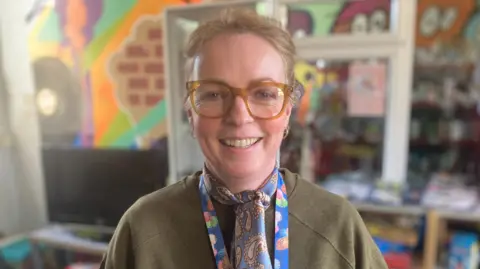 DJ McLaren/BBC Anna Foster, a woman who is standing inside and looking directly at the camera and smiling. She is wearing a green khaki jumper and a blue paisley scarf. She is wearing brown glasses.