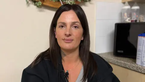 Krystal Ogier, a middle-aged woman, wearing a neutral expression. She is wearing a black hoodie with a grey T-shirt underneath. She has a tattoo on her neck and long, dark brown hair.