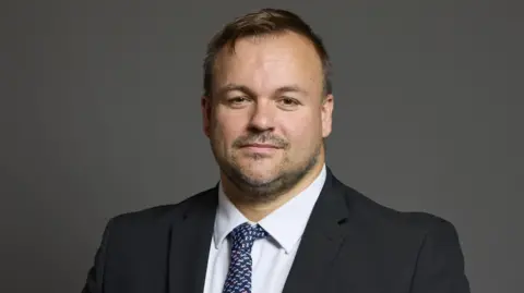 UK Government Terry Jermy is wearing a black suit with a white shirt and a blue tie. 
He has short, brown hair and a short beard and moustache.
He is standing in front of a grey background for his official portrait from the House of Commons.