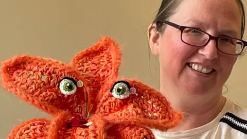 Rugby Yarn Bombers A woman wearing glasses with her hair tied back smiles at the camera. Next to her is an orange knitted flower which has crafted eyes.