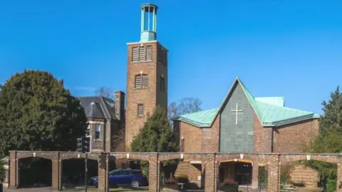 A 1950s church is shown under clear blue skies with trees to its sides. It has a faded blue roof and a cross on its exterior with a tall tower and arched walls.