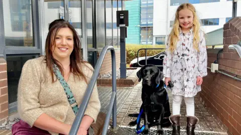 A woman is sat beside some outdoor steps. A black labrador is sat and a girl is stood on the steps. The woman has long dark hair, a gold jumper, maroon trousers and a green bag with a long strap across her body. The dog is wearing a blue collar, lead and harness. The girl has long blonde hair, a white dress printed with images of dogs, white leggings and furry brown boots. There is a grey-fronted building with an intercom, a grey handrail and a brick wall to the right. There is a black car and green college building in the background.