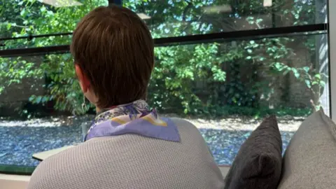 Lydia Dowling Ranera/BBC A woman is pictured looking out of a window. You can only see the back of her head. She has short, dark hair and is wearing a beige jumper and has a patterned purple scarf around her neck