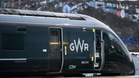 Image of a GWR train pulling into Bristol station. The dark green train takes up most of the image, with the Totterdown houses seen blurred in the background. 