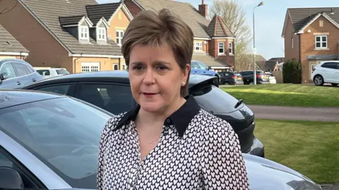 Nicola Sturgeon has short brown hair and is wearing a black and white top. She is standing on the driveway of her house with a grey car and a black car in the background 