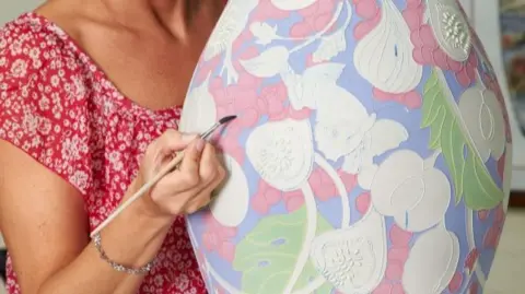 A woman decorates a pottery vase with a flower design on it. We do not see her head but she wears a red dress with white flowers on it and holds a paintbrush with the tip on the vase. Parts of the vase are not painted but some have red, green and blue paint on already.
